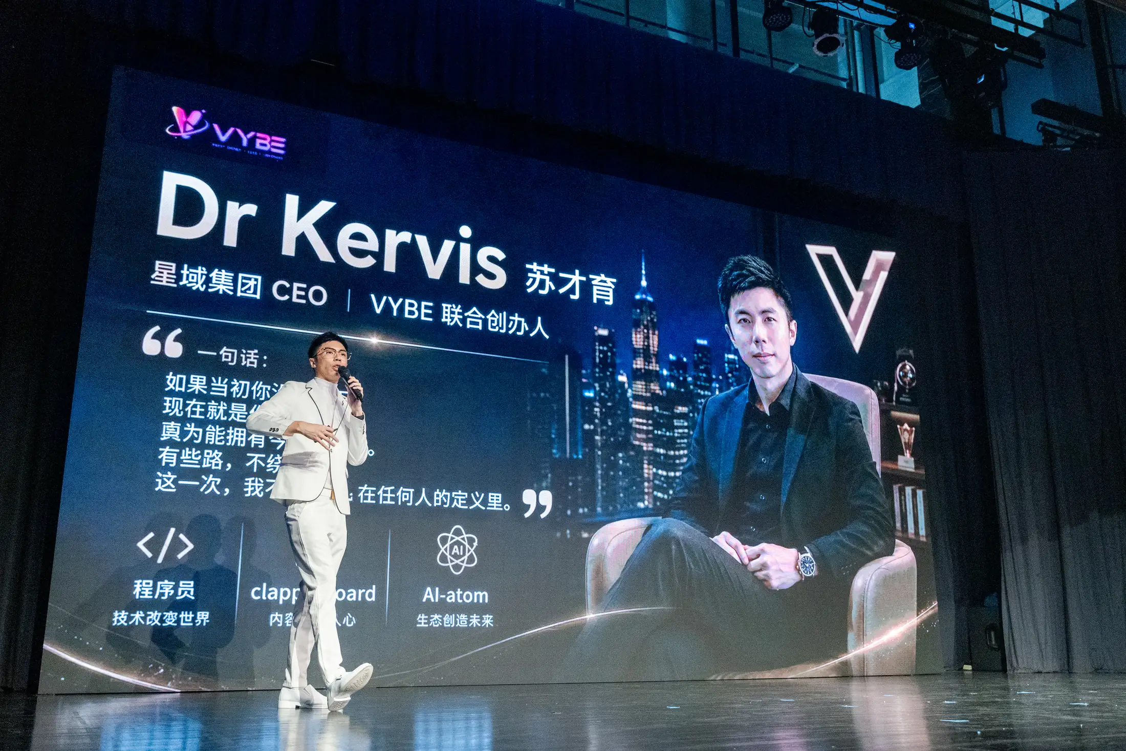 苏才育博士身穿白色西装、手持麦克风在舞台上演讲，身后大屏显示他的姓名与 VYBE 标识